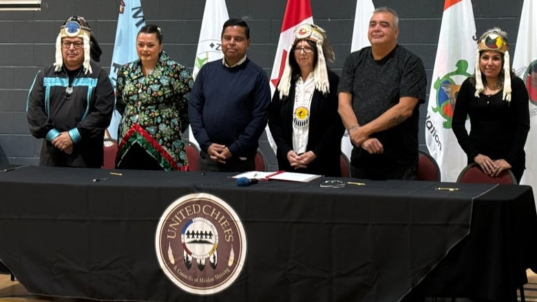 Six people, five in regalia, stand behind a long black table with flags behind them.