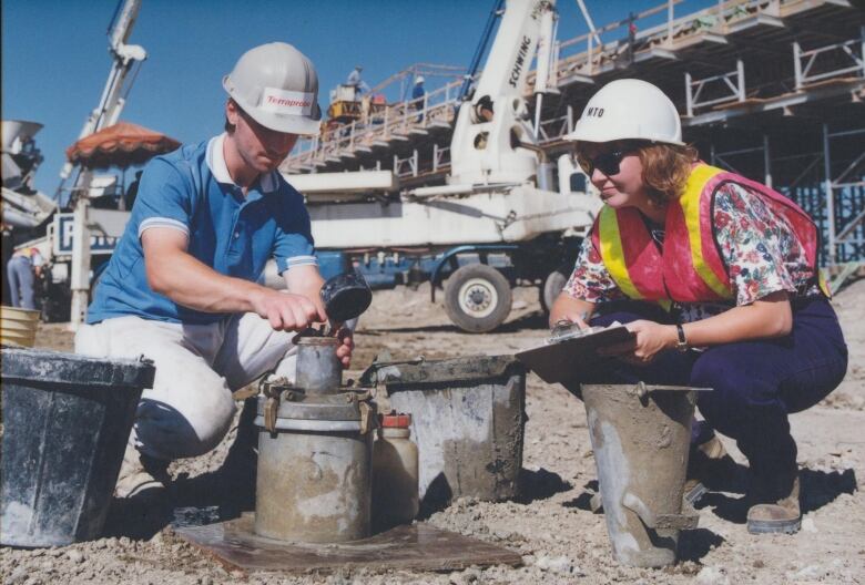 Two construction workers crouch on a job site. 