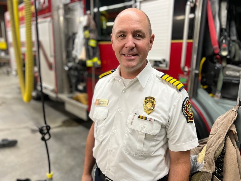 Man stands inside the garage of a firehall with a firetruck behind him. He is wearing a white uniform style shirt. 