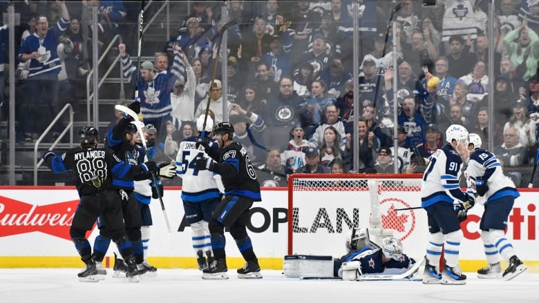 A group of men on black hockey jerseys hold sticks up on the ice, while a group of men on white jerseys stand some on the ice.