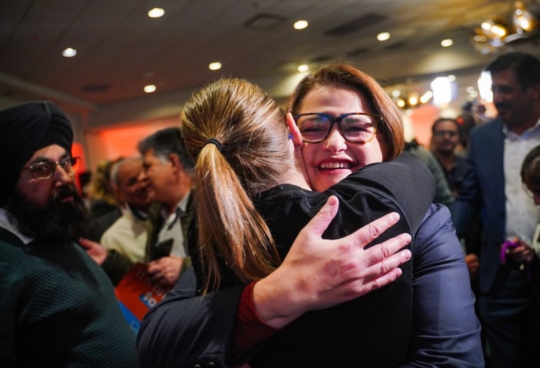 A woman in glasses enbaces another woman in a corwded room.