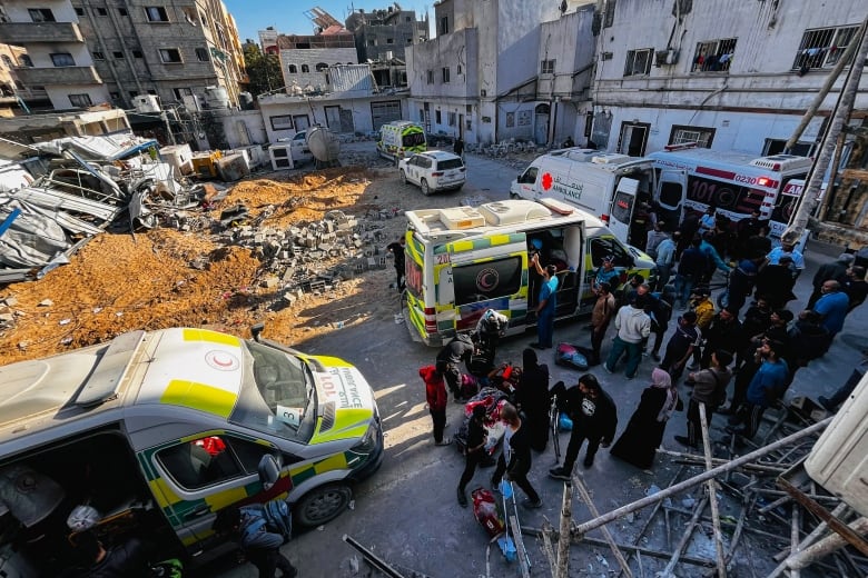 Dozens of people are seen near an ambulance and other vehicles in an urban setting.