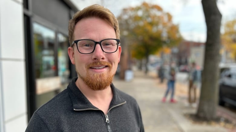 A man with red hair, glasses and a beard, smiles at the camera outside on a sidewalk with fall leaves in the background. 