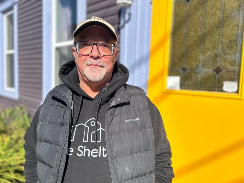 A man with glasses, a hoodie sweater, a vest and a ball cap looks seriously at the camera with a purple house and bright yellow door behind him.