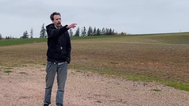 A man in a black hoodie stands on a gravel driveway in a rural area pointing towards the road. 