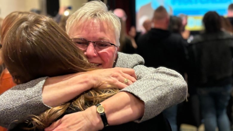 A woman in a grey suit jacket hugs a woman in black.