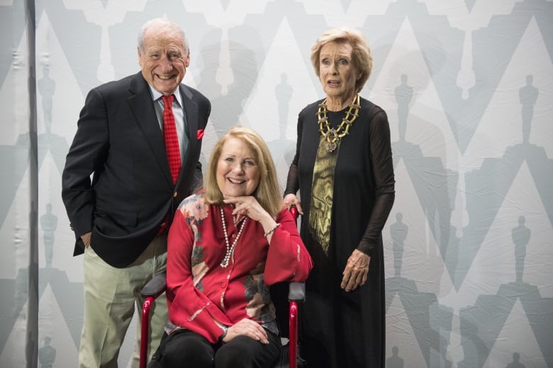 A blonde hair woman sits in a wheelchair with a man standing to her right and another woman to her left. 