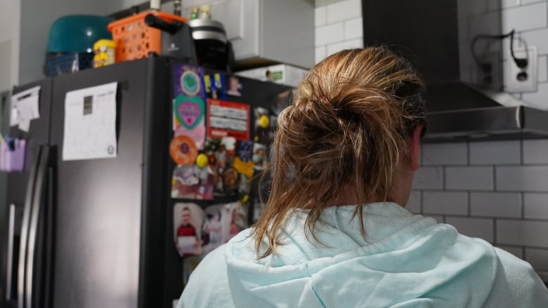 A woman in a blue sweatshirt with her hair in a messy bun stands in front of a stove. The side of the fridge next to her is covered in photos, magnets and calendars.