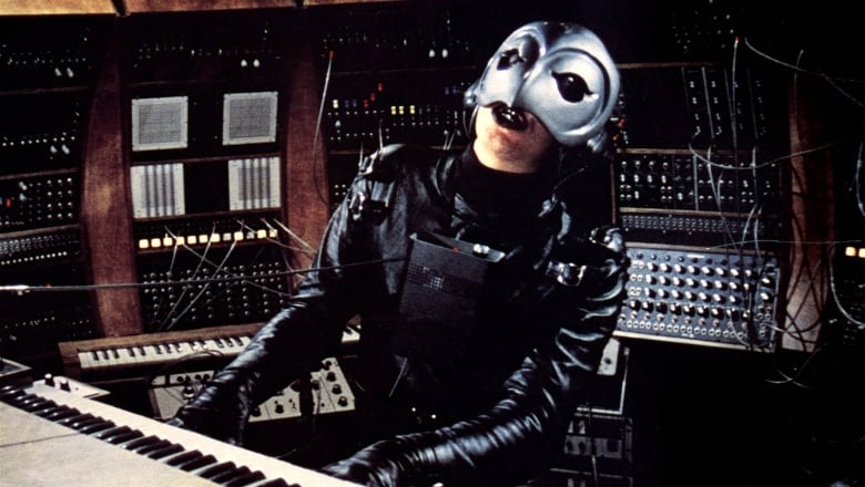 A man wearing a black leather suit and a silver mask that covers half his face plays a keyboard, surrounded by synthesizers and other musical equipment.