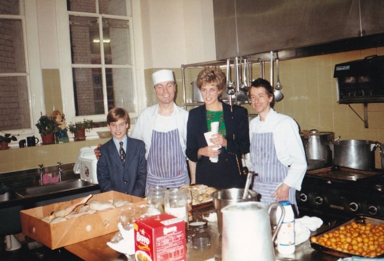 Three adults and a child stand behind trays and bulk packages of food in a large kitchen.