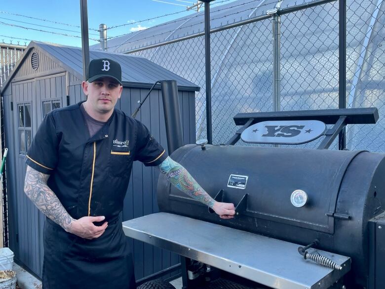 A man in a black ballcap and black chef wear stands beside a large metal cooker.