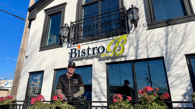 A man in an all black outfit stands in front of a white building with giant lettering over his head, reading Bistro on B.