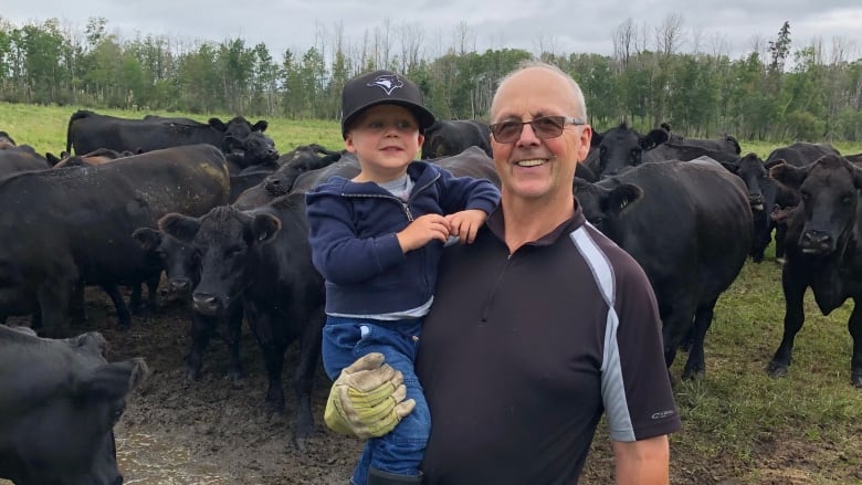 An older man with glasses holds a young child in one arm. There are cattle in the background.