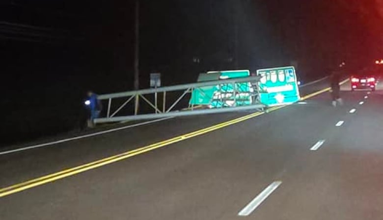 An aluminum structure with green highway signs is shown laying damaged on a road.