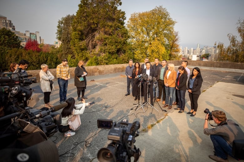 A group of politicians stands together in front of a microphone on a concrete plaza as cameras film them and journalists listen.