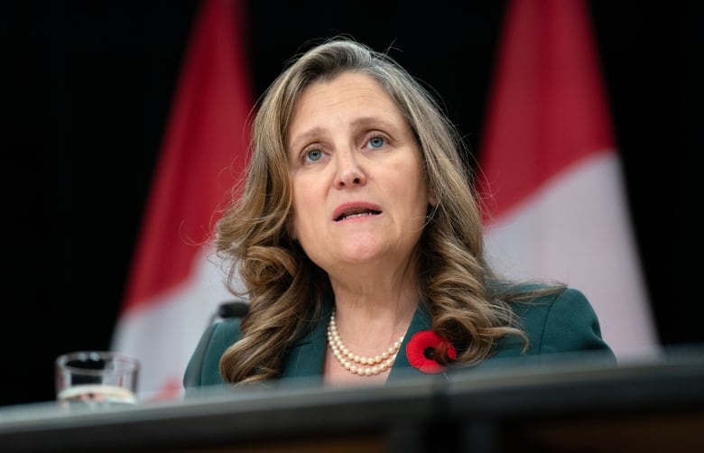 A woman in a green blazer speaks in front of a row of Canadian flags.