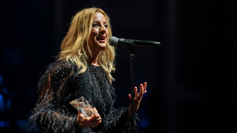 A woman with light coloured hair just a few inches past her shoulder and wearing a dress speaks on a stage while holding what appears to be an award.