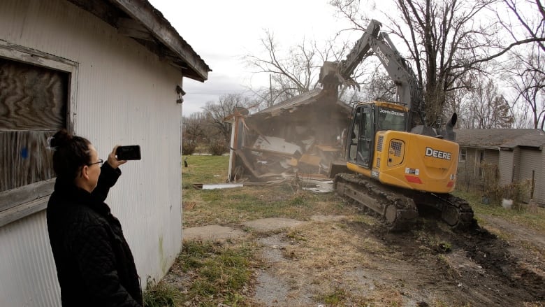 Woman holds phone to take photo of an excavator demolishing a building.