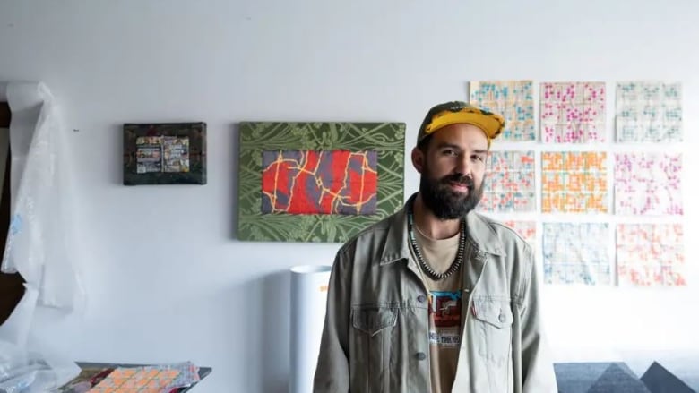 A young bearded man wearing a yellow cap with a beaded brim stands in front of a white wall with graphic beaded artworks on the wall behind him.