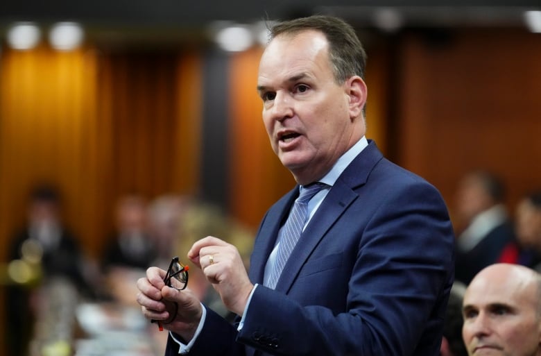 A minister stands in the House of Commons, wearing a navy suit and grasping a pair of reading glasses. 