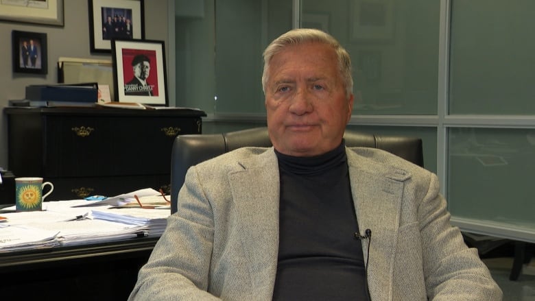 A man wearing a black shirt and a light grey blazer sits in his office.