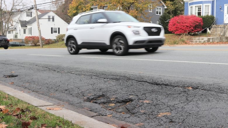 Some damage is seen on a road with a car going by. Houses are in the background and it's clearly fall. 