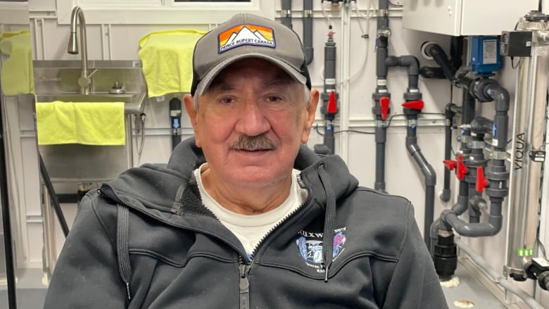 Older man in a grey hat and black zip up sits in a building filled with pipes and dials.