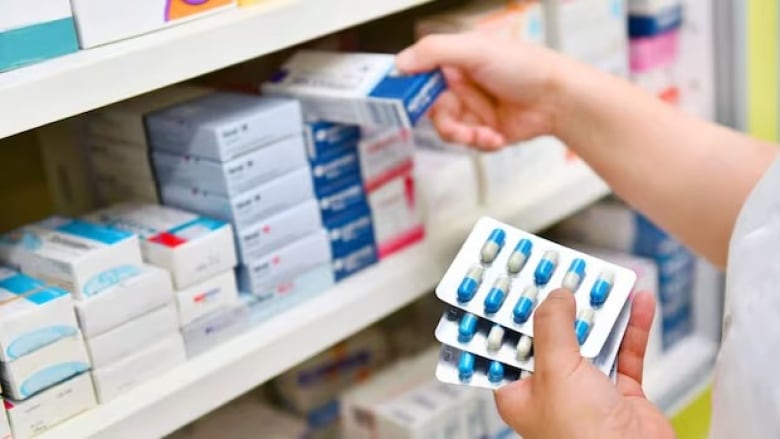 A pharmacist's hand is seen holding birth control pills. A shelf of more birth control is in the background.