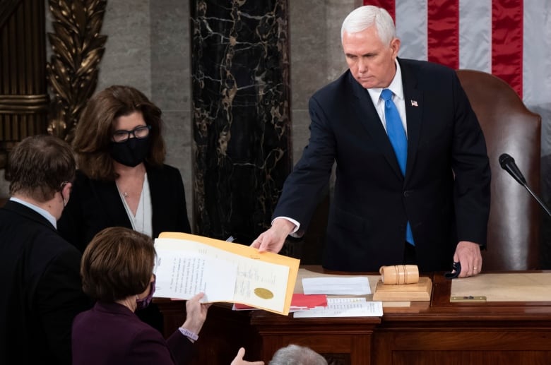 A cleanshaven white haired man in a suit and tie reaches out with his right hand to receive a signed document from a woman. The handoff is occurring in what looks to be a legislative chamber. 