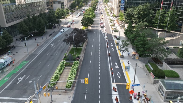 An aerial shot of University Avenue in downtown Toronto on a summer day. The avenue is mostly empty. There are 3 lanes for cars on each side and a bike lane on each side, surrounded by buildings 