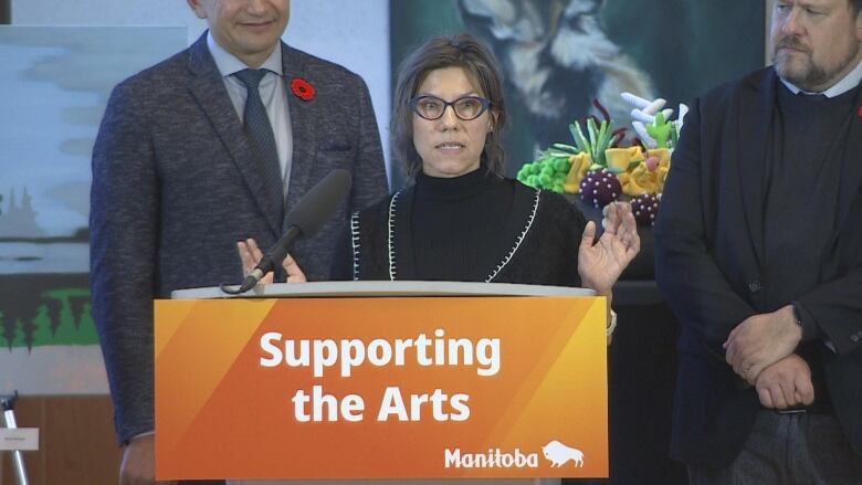 A woman with short brown hair, wearing a black shirt, speaks at a podium that says 'Supporting the Arts.'