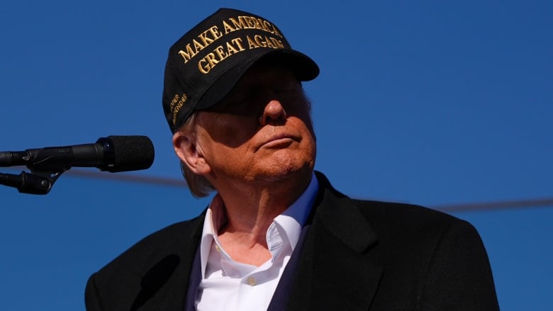 A man standing at a microphone looks to his left. He's wearing a black baseball cap, his face in shadow. 