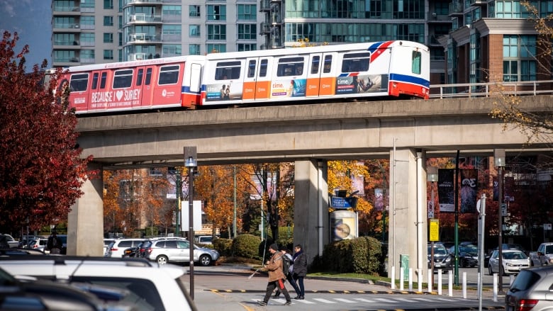 A SkyTrain passes above a busy intersection, with cars and pedestrians visible.