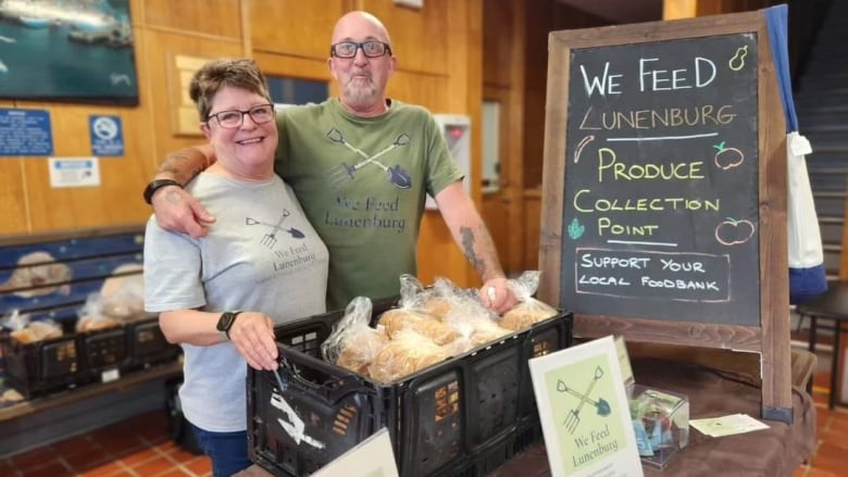 A couple side-hug and grab a basket with food next to a sign that reads 