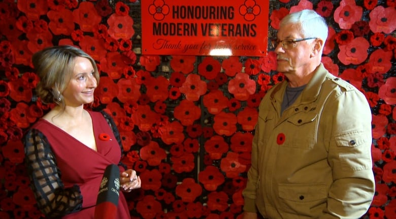 A man and woman stand in front of a wall of a couple hundred hand-made poppies. They are bright red and black. 