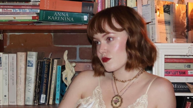 A woman with short red hair sitting sideways in front of a bookshelf wearing a beige satin lingerie set