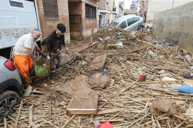 Two men carry a bucket of mud in a street covered in flood debris.