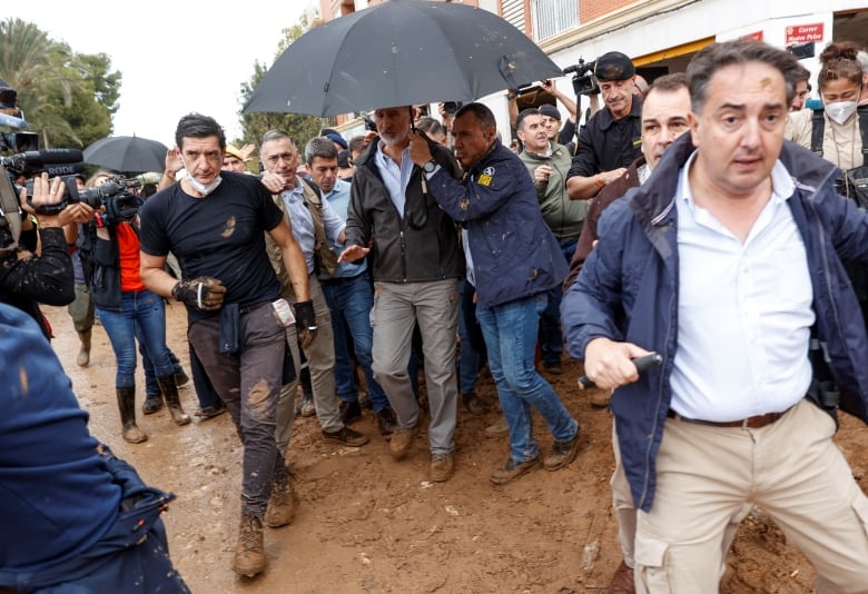 The king of Spain walks under an umbrella while people around him are pelted with mud.