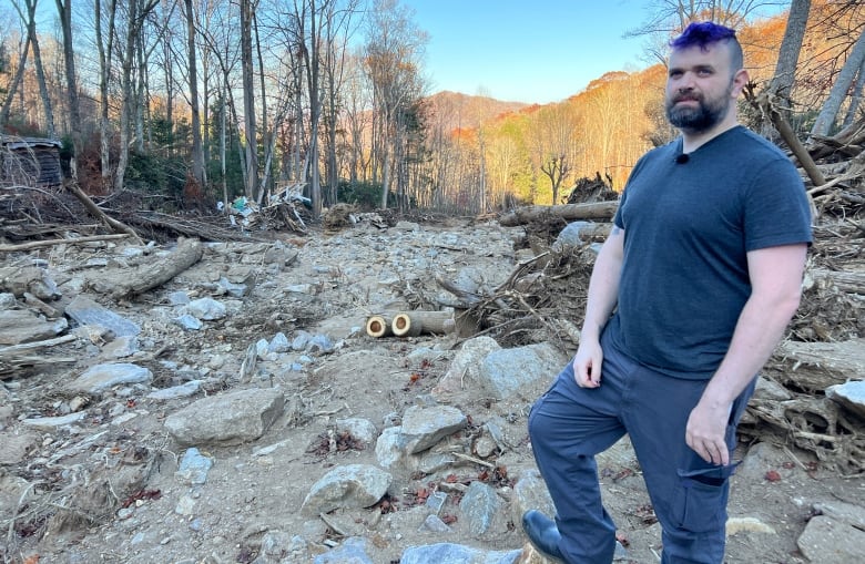 A person looks at the camera while standing amid debris and fallen branches.