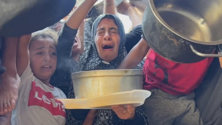 A woman holds out a pot as she sobs in a crowd of children and other people.