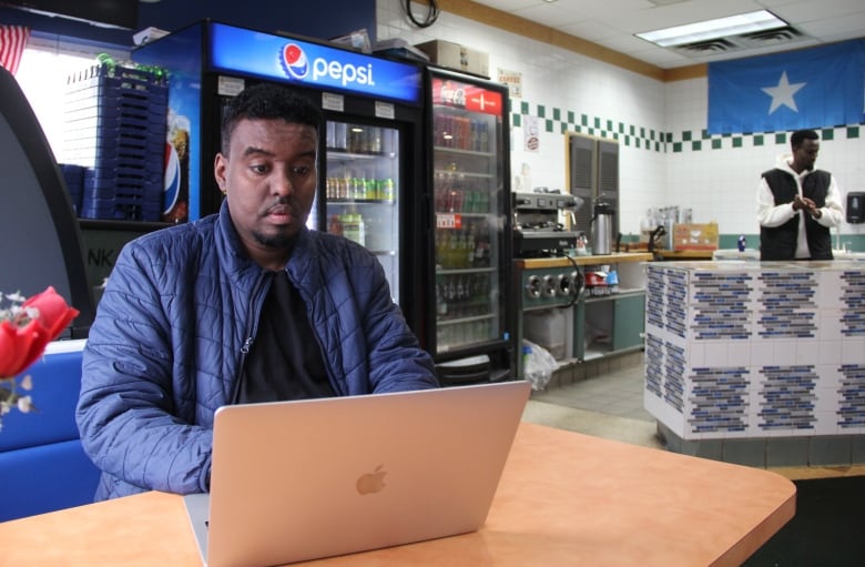 A man in a blue jacket and a black shirt underneath is seated at a table at a restaurant, typing on his laptop, while behind the counter a man has his hands together.