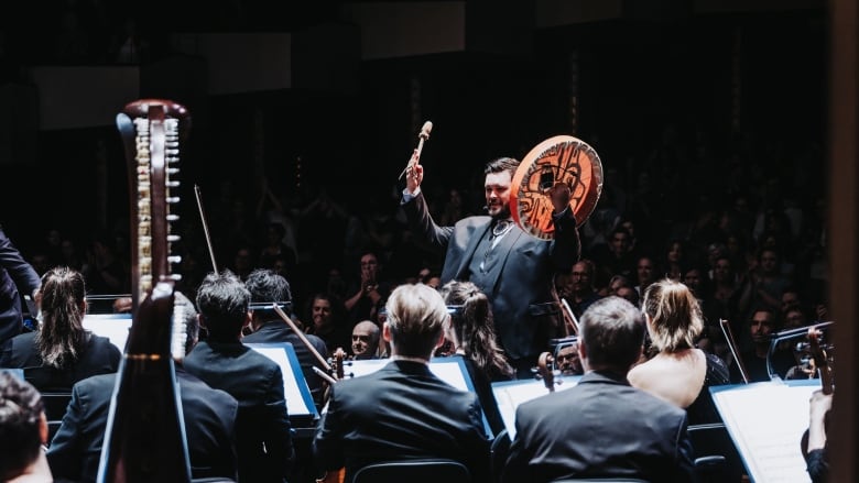 A man holds a drum in front of an orchestra