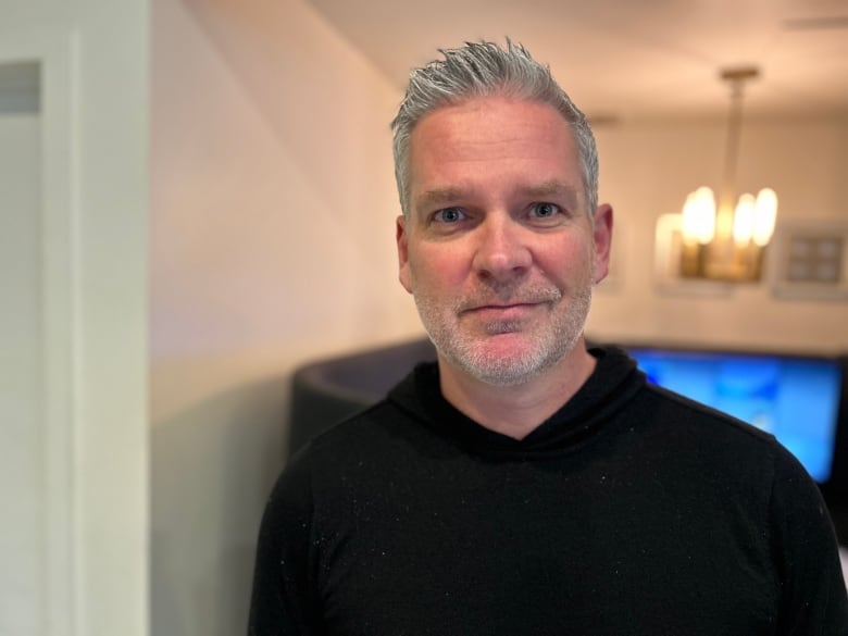 A man with salt and pepper hair and a beard smiles at the camera, while wearing a black sweater. 