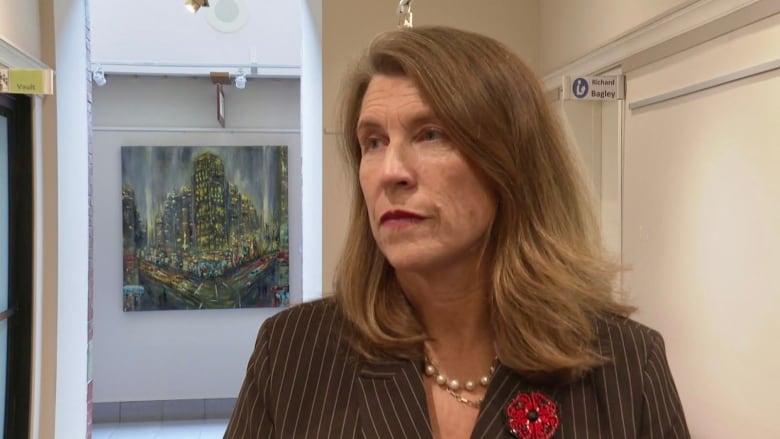 A woman with brown shoulder length hair looks to her right, with closed mouth, wearing a poppy and a brown blazer.