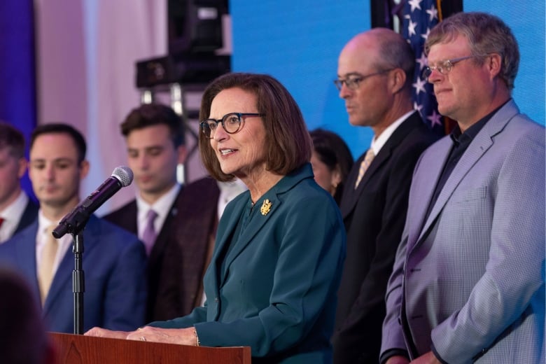 A woman speaks at a podium, flanked by men in suits.