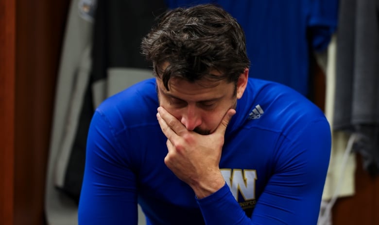 A football player in a blue shirt looks solemn, his chin resting in his hand.