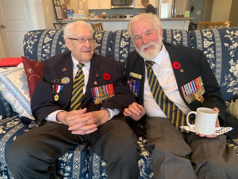 Two men in suits with medals on sit side by side on a sofa, smiling. 