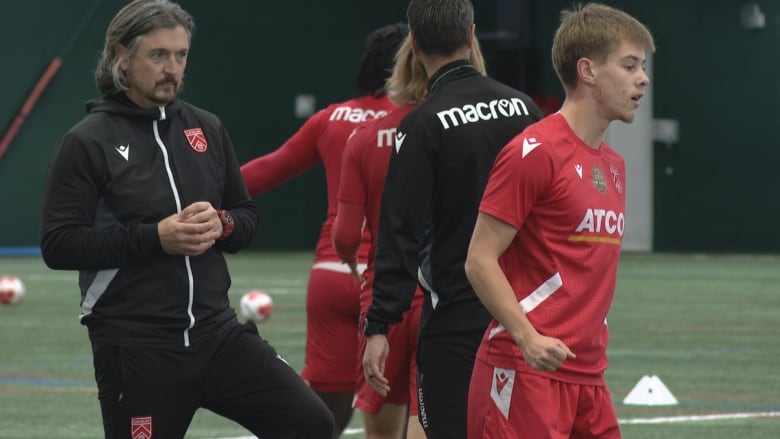 A man wearing black is pictured on the left watching a group of people wearing red warming up ahead of a soccer practice.