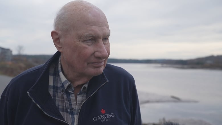 A bald man stands oputside into front of a river whearing a jackewt that says Ganong.