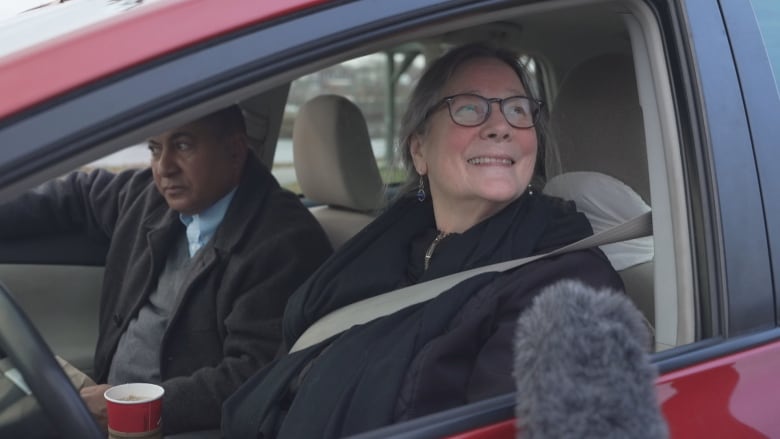 A woman sits behinmd the wheel of a red vehicle. Her head is turned toward a large fuzzy microphone being held beside her rolled down window.